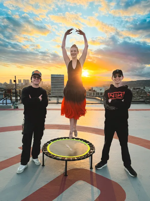 A ballerina mid-jump on a trampoline at sunset, flanked by two smiling crew members