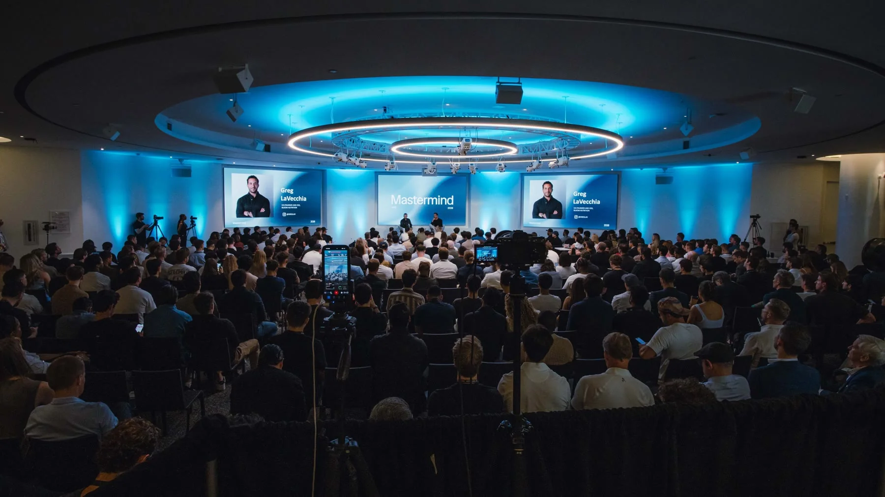 The NYC Mastermind room seen from the back, a full audience facing the stage under a ring of blue light